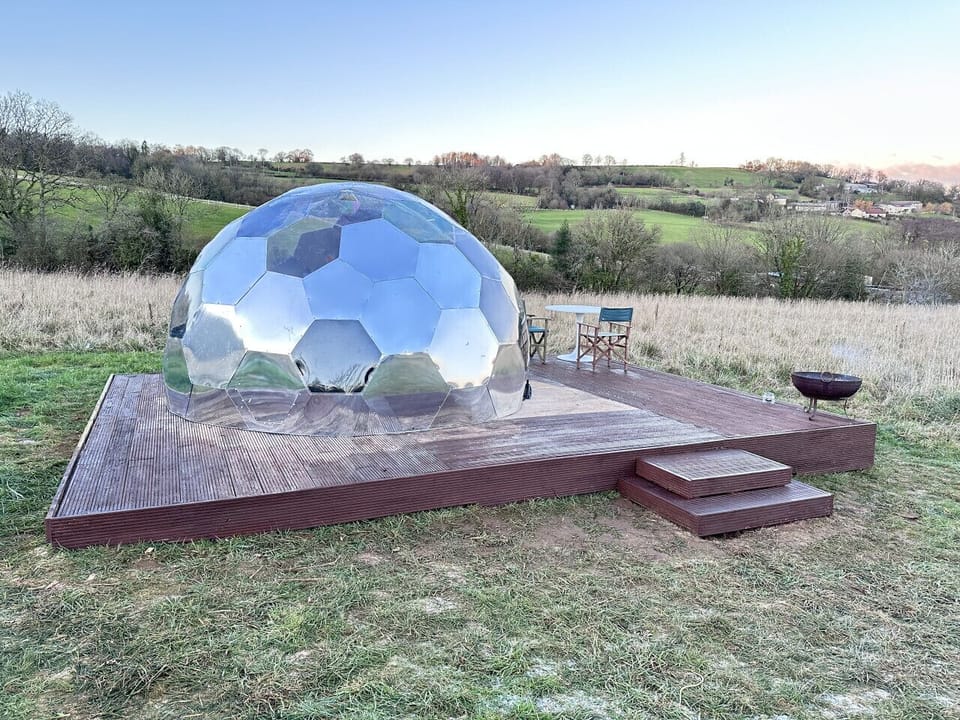 The dome is set on a wooden platform in an open field with countryside views