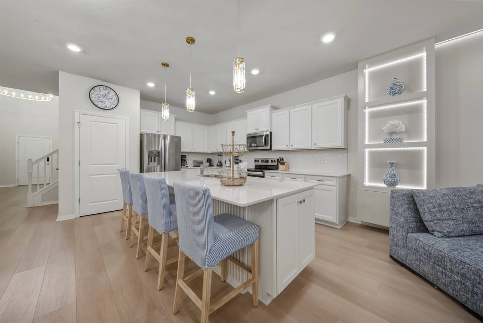 Welcome to a bright kitchen with sleek white cabinetry and a spacious island adorned with soft blue bar stools, ideal for casual dining or coffee mornings, while modern light fixtures enhance the inviting atmosphere.