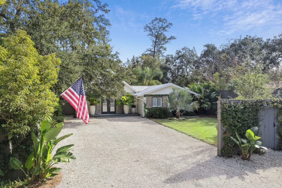 Charming property entrance with American flag welcomes guests through lush tropical landscaping and mature oak trees.