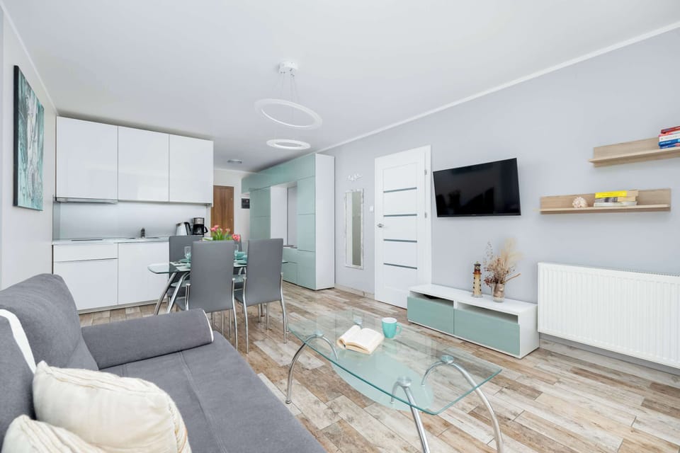 A living room with a wall-mounted TV and comfortable gray seating. The glass coffee table adds elegance to the bright open area.