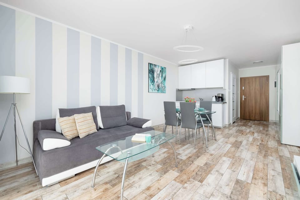 A bright living room with striped walls, a gray sofa set, and a glass coffee table. Natural light fills the space, giving it an airy and inviting feel.