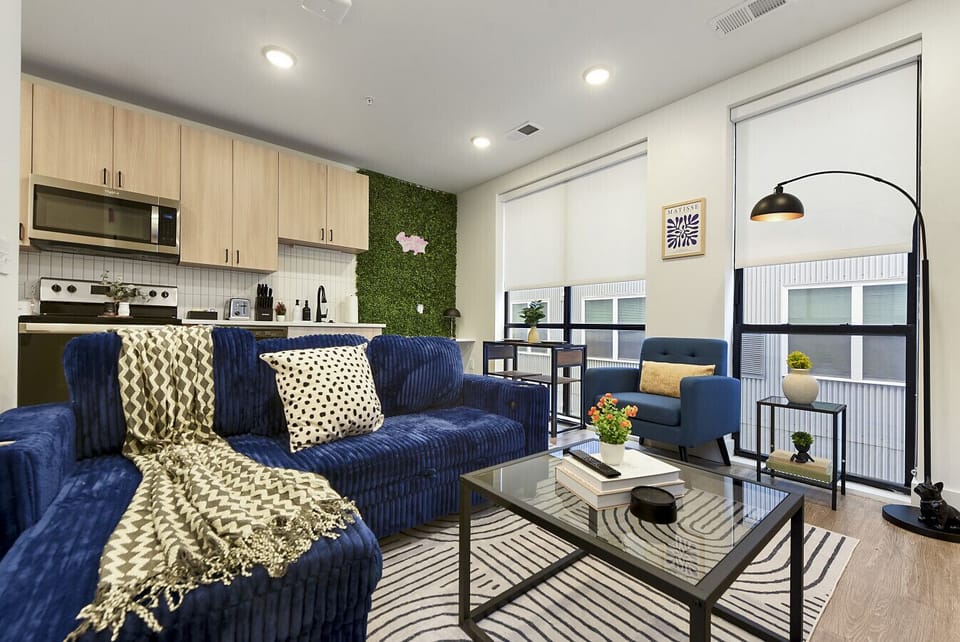 Bright, fully equipped kitchen with stainless steel appliances, dishwasher, coffee station, and seamless open flow to a comfortable blue sofa seating area accented by a lush green plant wall—great for easy stays.