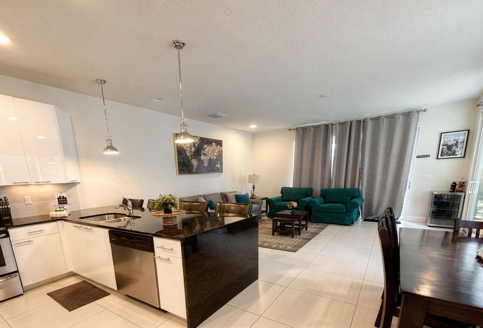 Enjoy a stylish kitchen with sleek white cabinets and dark countertops, ideal for cooking, flowing into a cozy living area with plush teal seating, all bathed in soft natural light for a warm and inviting atmosphere.