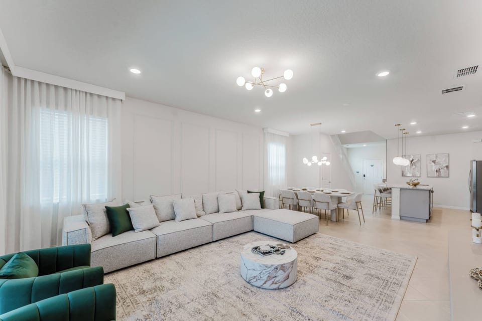 Enjoy the warmth of natural light in this spacious living area, where a cozy sectional invites you to relax amid soft neutral walls and a stylish chandelier that adds elegance, creating a welcoming atmosphere for all guests.