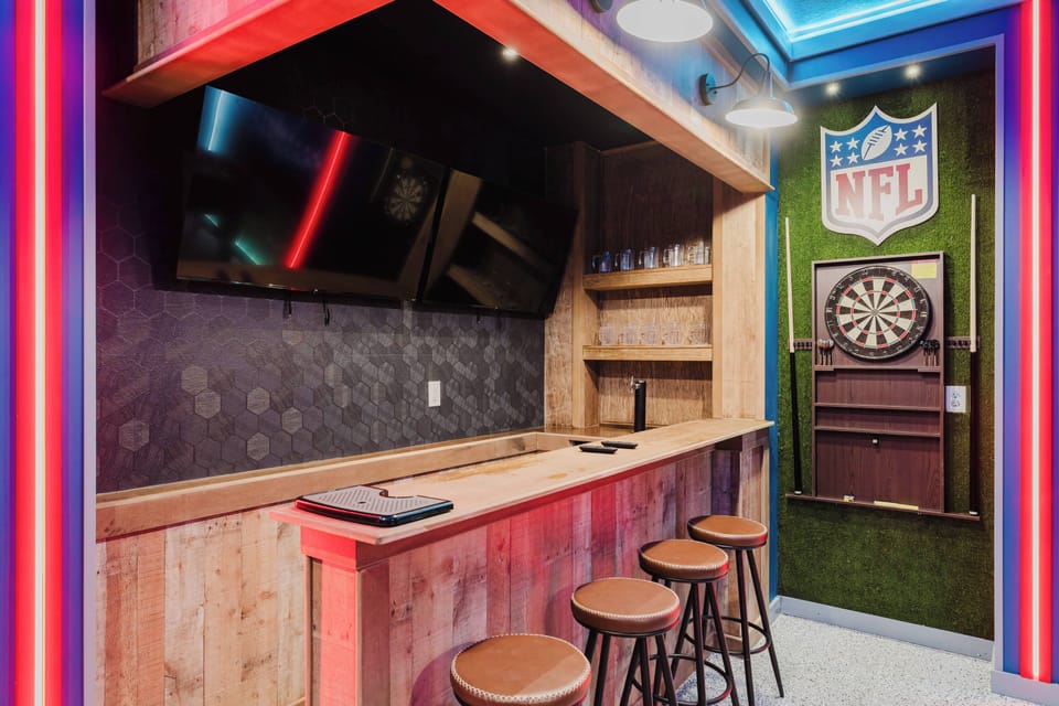 Experience the stylish game room with a beautifully crafted wooden bar and sleek stools, perfect for enjoying drinks while watching sports on the large TV. The vibrant neon lighting adds a fun touch to this inviting space.