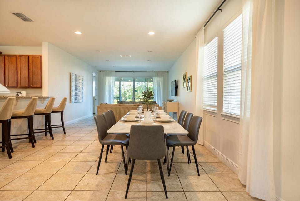 Sunlight streams through large windows, brightening the modern dining area with its chic table and gray chairs. The tasteful décor and warm tile floors create a welcoming space ideal for enjoying shared meals together.