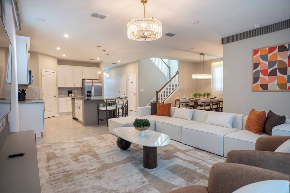 Experience a cozy living area with a plush white sectional sofa and warm earth-toned cushions, ideal for relaxation. A modern chandelier lights up the stylish coffee table, creating a seamless connection to the kitchen for easy.
