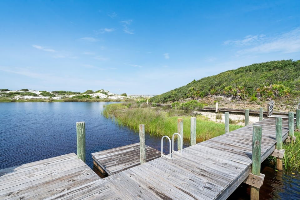 Pristine lake with wooden dock surrounded by rolling dunes and coastal vegetation under clear blue skies.