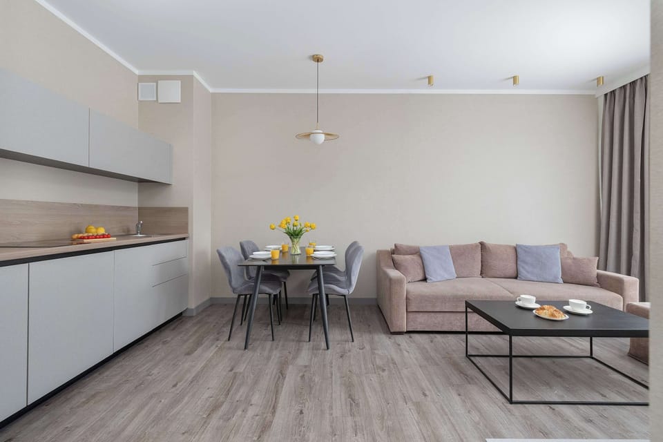 A bright open-plan living room with a sofa, dining table, and kitchenette. The neutral colors and large windows create a spacious and welcoming atmosphere.
