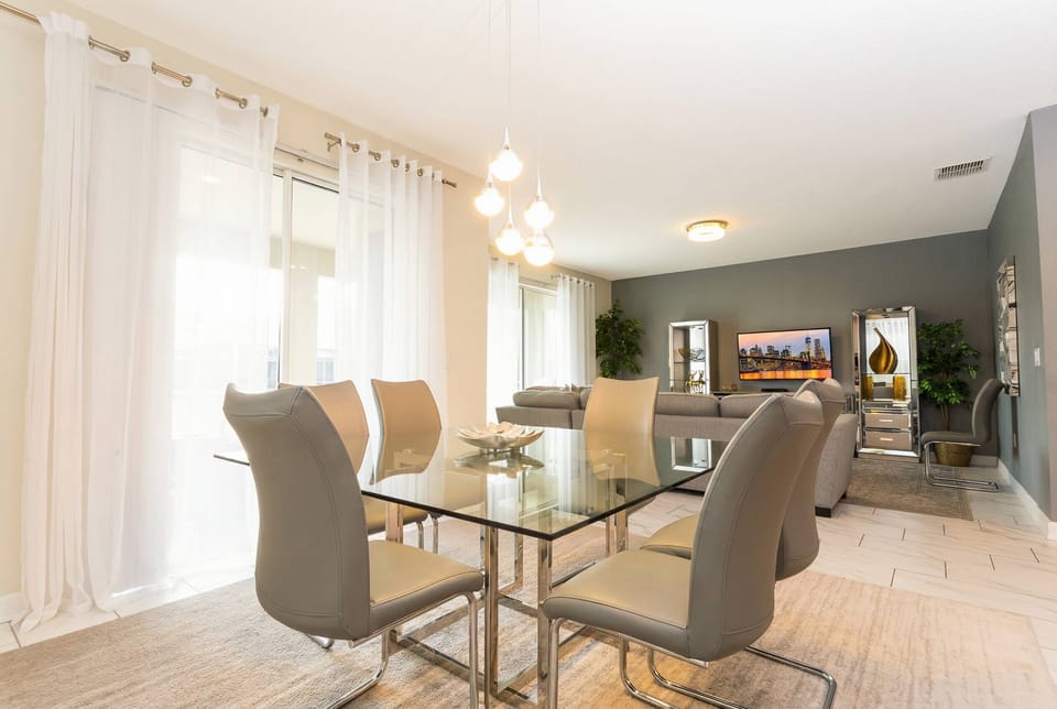 Gather in a bright dining area with a glass table, elegant chairs, pendant lights, soft white curtains, neutral decor, and an open view toward the stylish living room.
