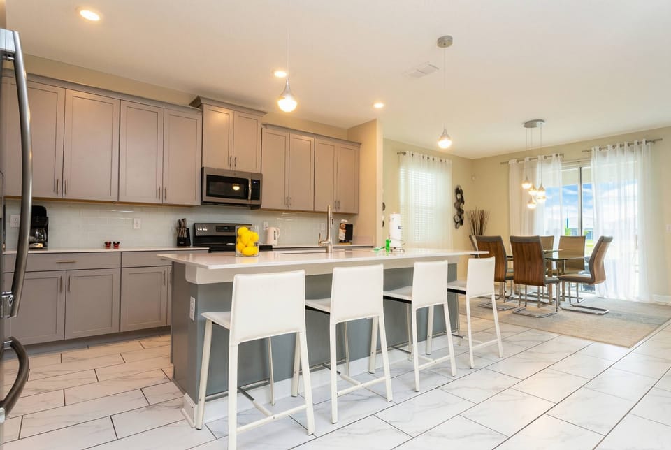 Cook and gather in a bright open kitchen with gray cabinetry, stainless steel appliances, a large island, white bar seating, pendant lights, and a connected dining area with soft curtains.