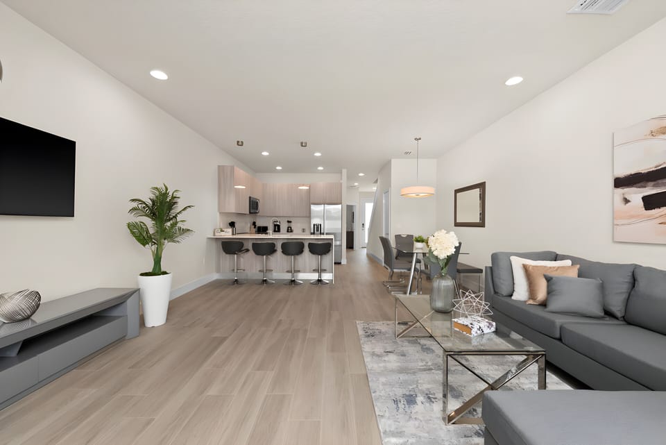 Welcome to a bright living room with a sleek gray sectional and stylish coffee table, perfect for relaxation. The open layout flows into a chic kitchen with modern bar stools and warm lighting, creating an inviting space for your.