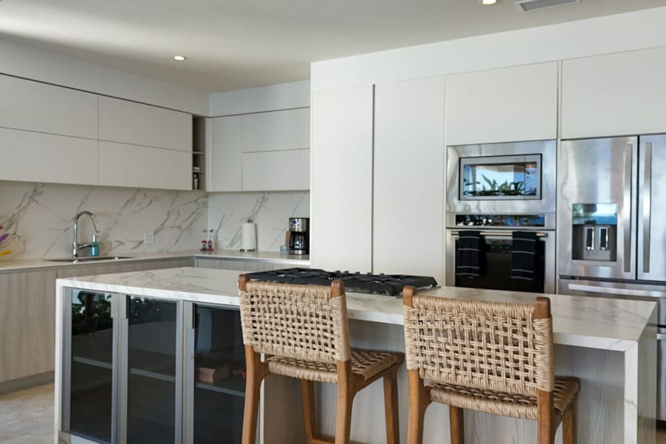 Modern, sleek kitchen featuring white cabinetry, marble countertops, and stainless steel appliances. The island includes a built-in gas stovetop and wine fridge, complemented by woven barstools for a stylish yet functional design.