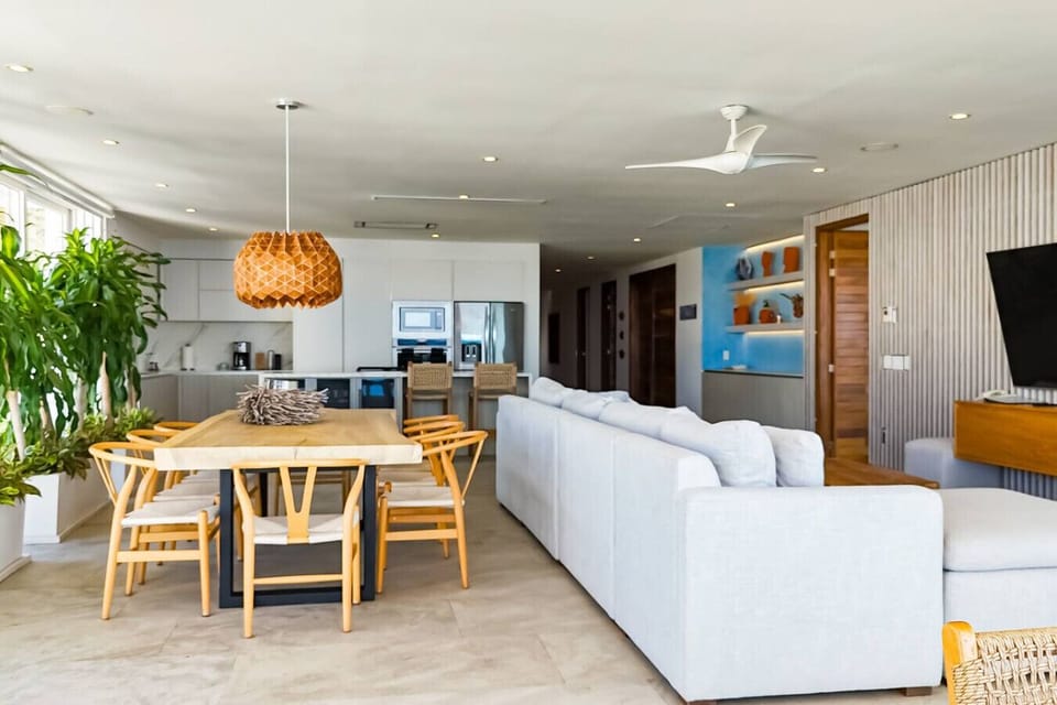 Spacious open-concept living area with a plush sectional sofa, natural light, and modern décor. Enjoy a seamless indoor-outdoor experience with floor-to-ceiling windows, lush greenery, and a fully equipped kitchen in the background for entertaining.
