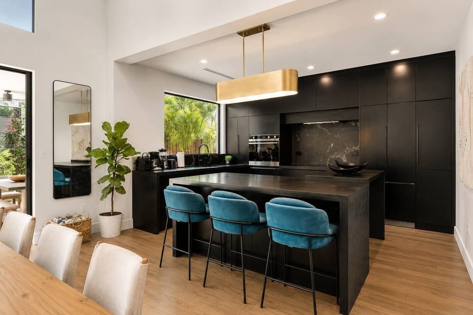 Prepare meals in the sleek, modern kitchen featuring black cabinetry, a dark marble-look backsplash, and a built-in oven. Three teal velvet barstools at the large black island provide a stylish spot for morning coffee or snacks.