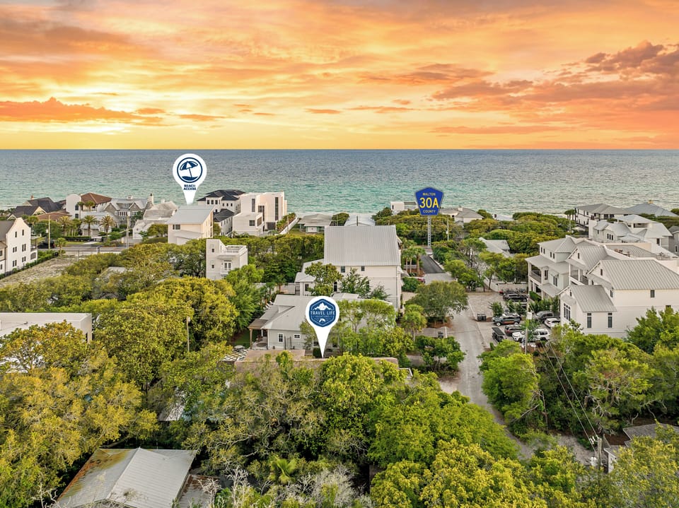 An aerial view captures a coastal neighborhood with beach homes nestled among lush greenery, just steps from the ocean at sunset.