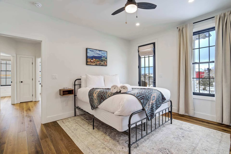 The primary bedroom features a comfortable bed and direct access to a private balcony through a glass door. Enjoy the convenience of a ceiling fan and bright natural light while you relax in this peaceful upper level space.