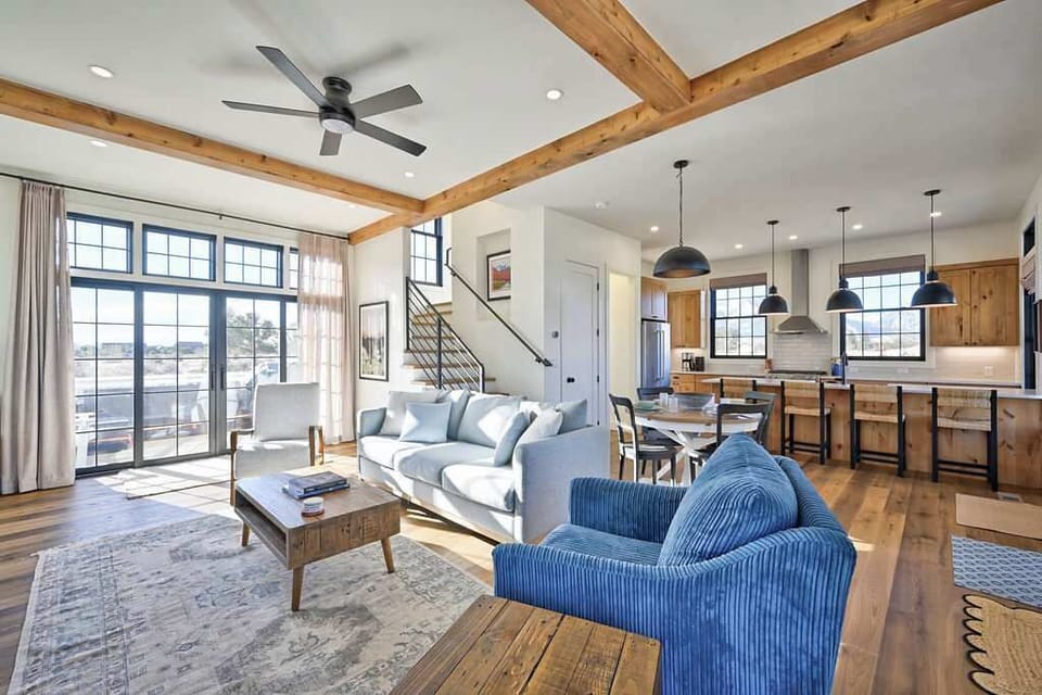 Inside, the living room offers a spacious feel with exposed wood beams, tall ceilings, and plush seating. The open layout connects to the kitchen while the modern black metal staircase leads you to the upper level bedrooms.