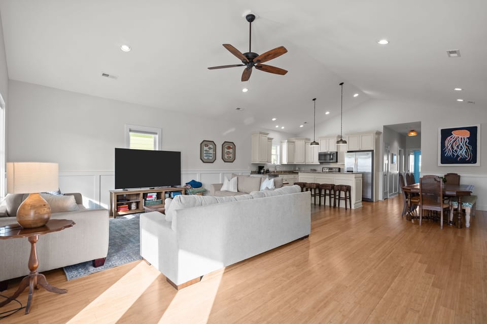 Airy living room with vaulted ceilings, large windows, and comfortable seating. Neutral tones, coastal artwork, and a spacious layout create a relaxing gathering space filled with natural light.