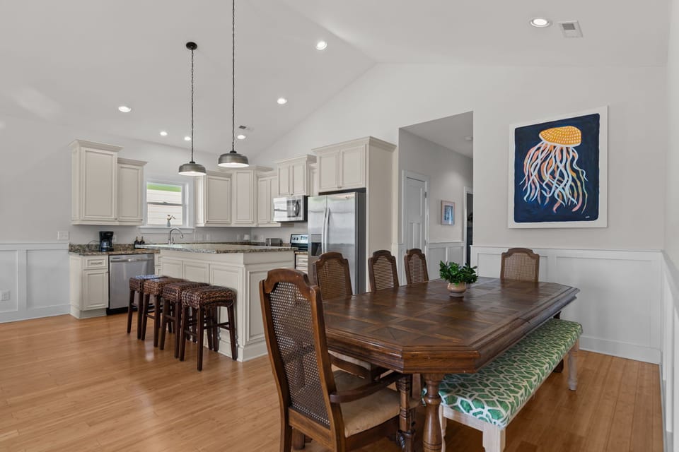 Spacious dining area featuring a wood table, wainscoting, and warm hardwood floors. Vaulted ceilings with recessed lighting flow seamlessly into the living space, creating a bright, airy coastal interior.