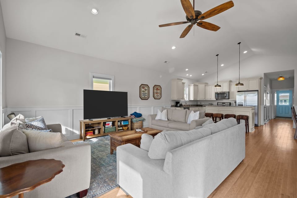 Airy living room with vaulted ceilings, large windows, and comfortable seating. Neutral tones, coastal artwork, and a spacious layout create a relaxing gathering space filled with natural light.