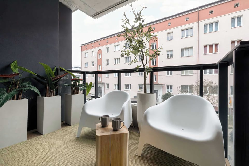 A balcony furnished with white chairs and a small round table. Potted plants line the railing, adding greenery and privacy. Surrounding residential buildings are visible in the background.