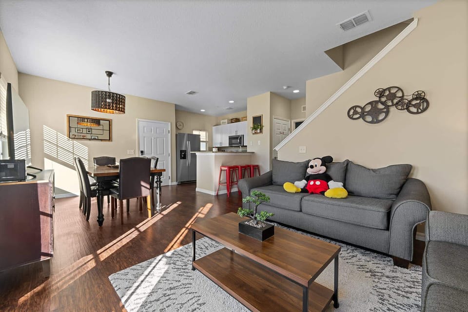 Sunlit living and dining area with modern furnishings and a playful mickey mouse plush on the sofa.