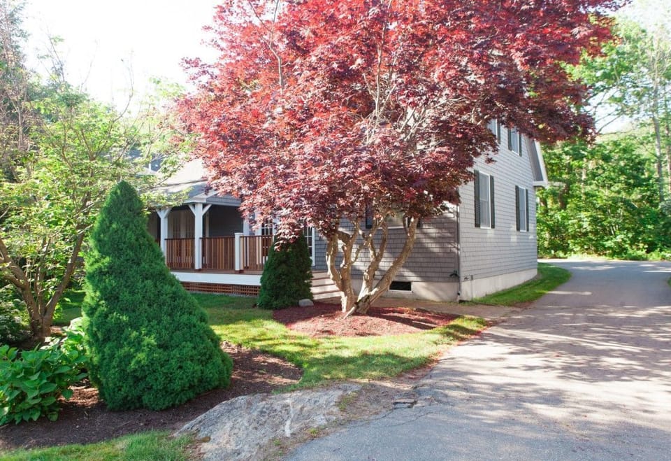 The cottage stands gracefully amidst lush greenery and vibrant landscaping, showcasing its stylish façade and welcoming entrance in the scenic Kennebunkport area.