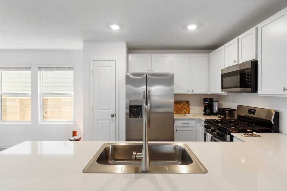 Bright kitchen and dining area featuring stainless-steel appliances, wide countertops, and a modern round table overlooking the backyard. The open layout flows into the living room, where a 55” TV creates the perfect place to relax after meals.