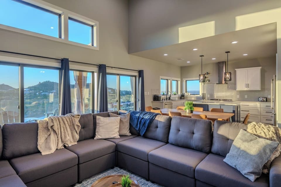 Spacious open-concept living room with oversized sectional, large windows flooding the space with natural sunlight, and seamless flow into the modern kitchen and dining area.
