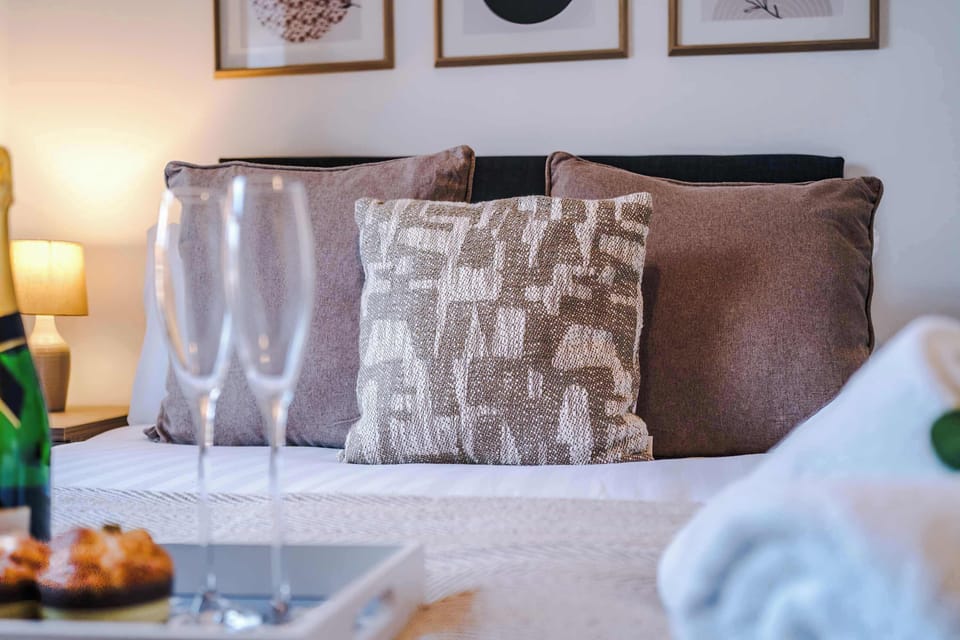 Bright and welcoming bed area featuring the upholstered headboard, decorative cushions, bedside table with lamp, and neatly presented bedding.