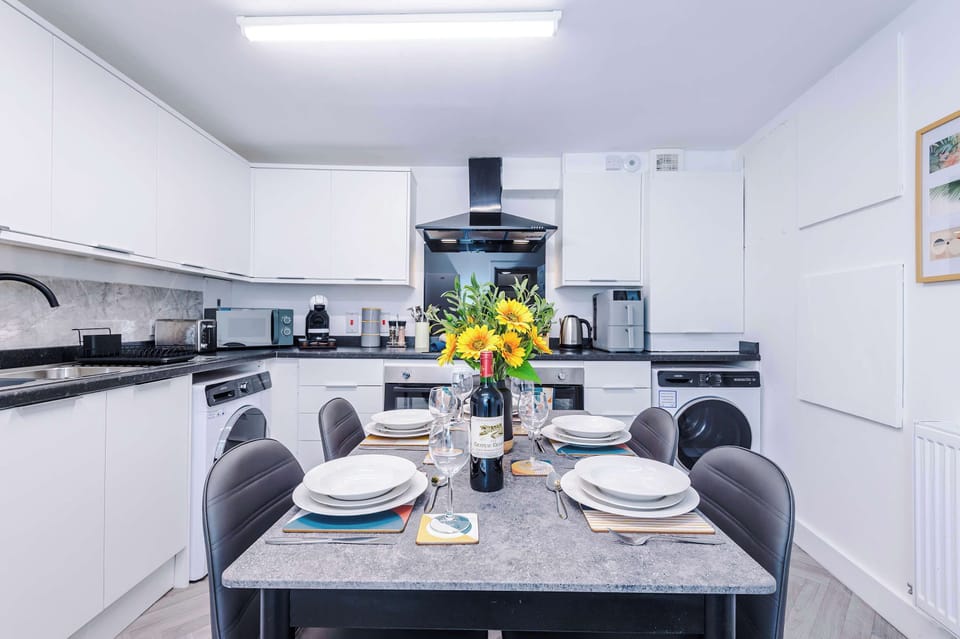A spacious, fully fitted kitchen featuring white cabinetry, integrated oven and hob with extractor, washing machine, and countertop appliances. The layout offers ample storage and worktop space, with a dining table ideal for everyday use.