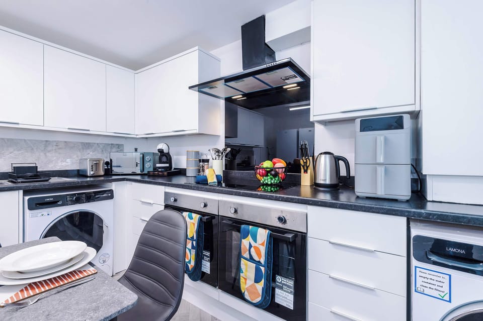 Bright, modern kitchen  work area featuring the integrated oven, hob with extractor, washing machine, kettle, and countertop appliances. Finished with white units and dark worktops for a clean, modern look.