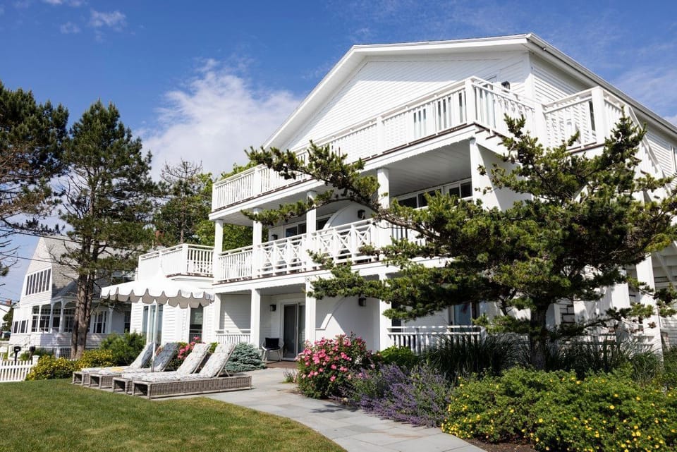 The pristine exterior showcases charming coastal architecture, surrounded by lush gardens and inviting lounge chairs overlooking the vibrant beach.