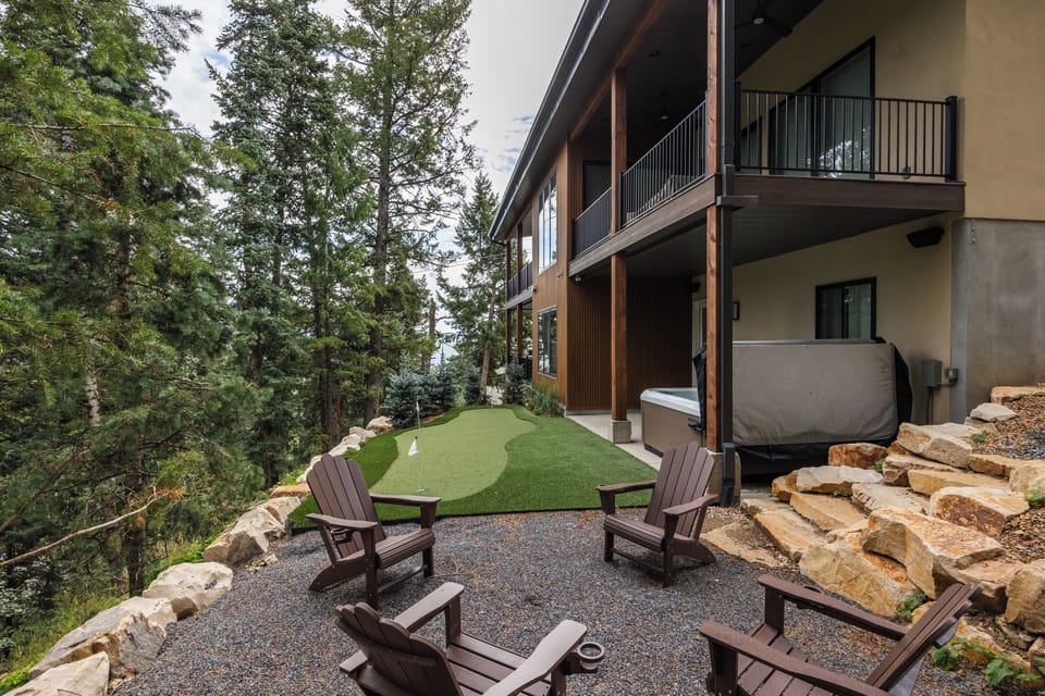 Unwind in the beautifully landscaped backyard featuring a hot tub, putting green, fire pit, and multiple seating areas surrounded by mature pine trees.
