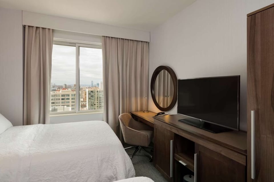 Guest room with desk, TV, and city view through large window