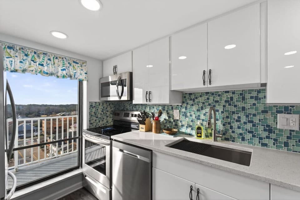 Modern, fully equipped kitchen with white cabinetry, stainless steel appliances, and a colorful tile backsplash. Sliding doors open to a balcony, filling the space with natural light and fresh air.