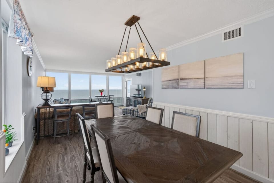Spacious dining area with a large table, elegant lighting, and coastal artwork. Open layout leads into a bright living space with ocean views.