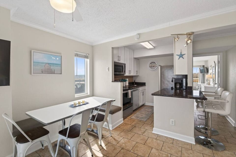 Bright dining area and fully equipped kitchen with modern appliances and coastal accents. Open layout with natural light, bar seating, and comfortable dining space.