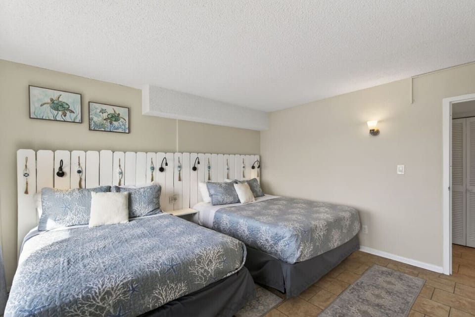 Bright and inviting bedroom featuring two comfortable beds with soft coastal-patterned linens, a crisp white shiplap headboard, and charming beach-inspired artwork.