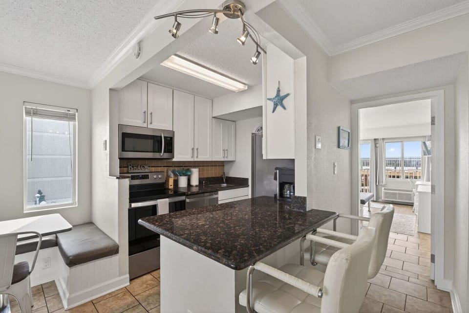 Modern kitchen with granite countertops, stainless steel appliances, and bar seating. Clean, bright space with coastal touches and an open layout that flows easily into the living area and ocean-view rooms beyond.