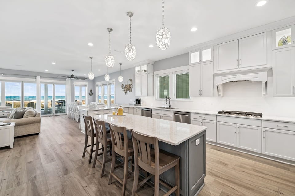 Your gourmet kitchen sparkles with white cabinetry, gleaming marble island, and crystal pendant lights. Ocean views through windows enhance every meal.
