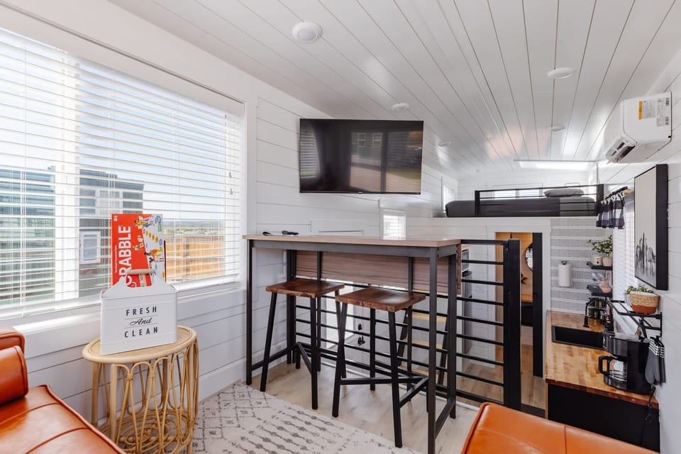 Modern loft-style living space featuring elevated dining area, smart TV, comfortable seating, and bright natural light. Perfect for relaxing, streaming movies, or enjoying a quiet evening inside your tiny home retreat.