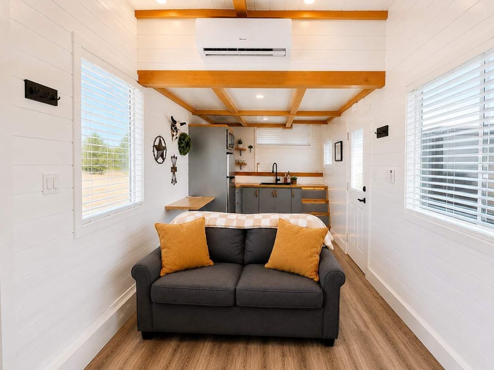 Open-concept tiny home living room with comfortable seating, Smart TV, electric fireplace, and modern kitchen nearby. Thoughtfully designed layout maximizes space while offering a bright, relaxing atmosphere and contemporary amenities.