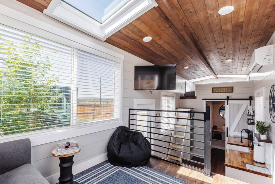 Elevated living area with sofa, bean bag chair, TV, large windows, and skylight bringing natural light into this tiny home near Zion National Park.