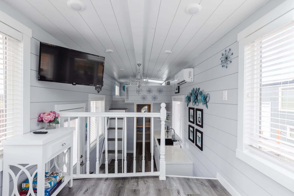 Light-filled loft living area with a TV, large windows, and an open layout overlooking the tiny home below.