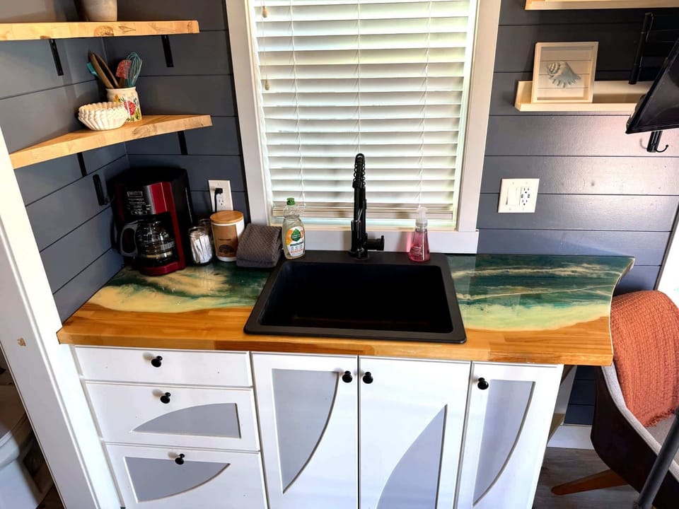 Custom ocean-inspired epoxy countertop, coffee station, sink, and open shelving bring unique coastal character to this fully equipped tiny home kitchen.