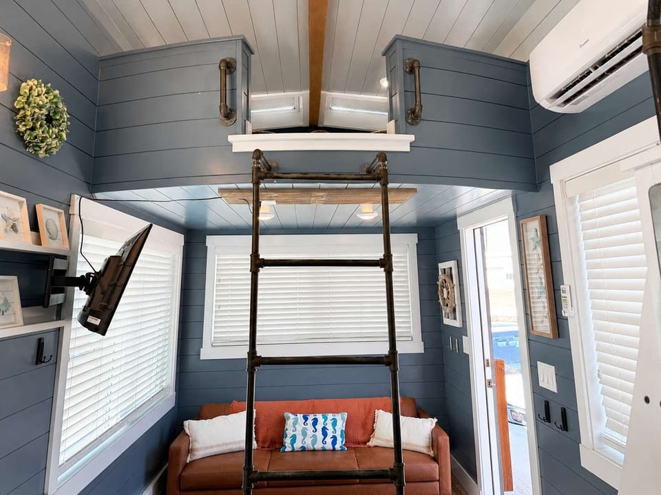 Open-concept tiny home living space featuring loft sleeping areas, ladder access, comfortable seating, and coastal-inspired décor. Bright windows and vaulted ceilings make the home feel spacious and inviting.