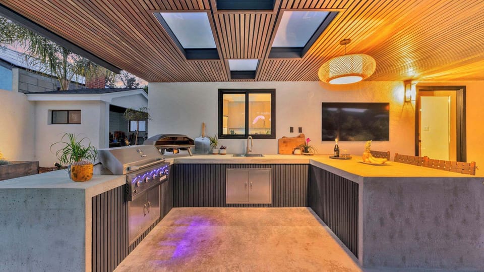 Full outdoor kitchen with U-shaped concrete counters, built-in grill, pizza oven, skylights, and pendant lighting — a chef-ready setup under the stars