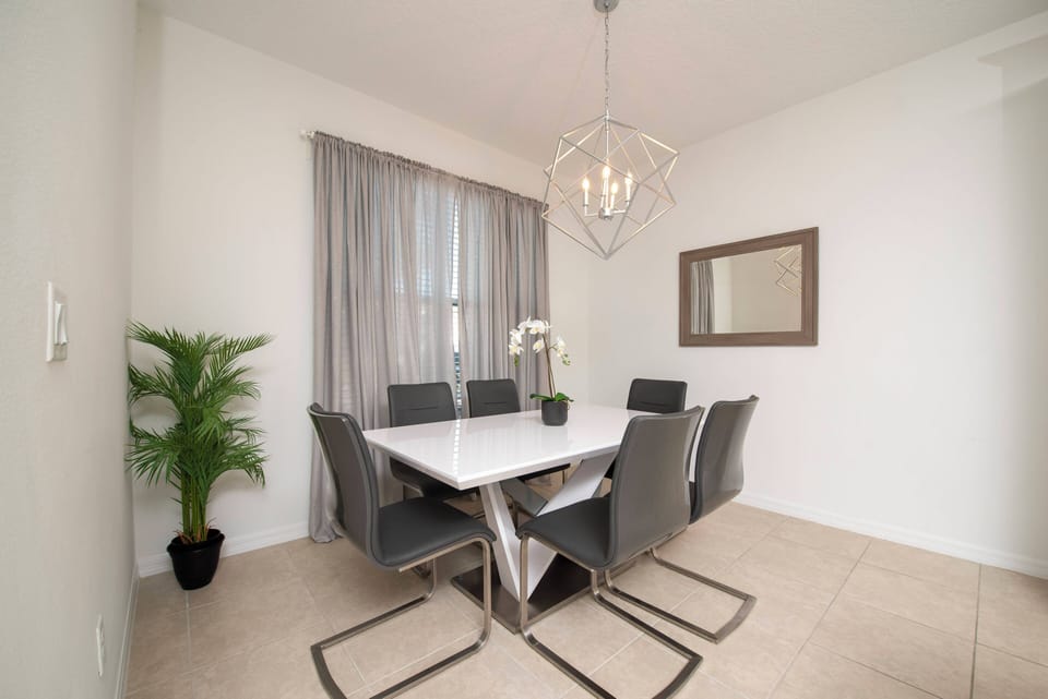 This bright dining area boasts a sleek white table with modern gray chairs, creating a stylish space for meals. A contemporary chandelier adds warmth, while lush greenery brings a refreshing touch to the atmosphere.